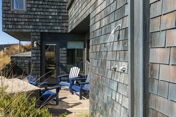 A modern building with a dark shingle exterior, a covered outdoor seating area with blue chairs, and a visible window. - At The Beach! Insanely Great Beach House, Ocean Views, Relaxation & Pickleball (Pajaro Dunes)