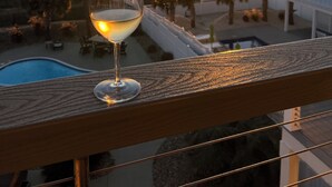 Ett glas vitt vin på en balkong med utsikt över en solnedgång, en pool och en byggnad med ett grönt tak.