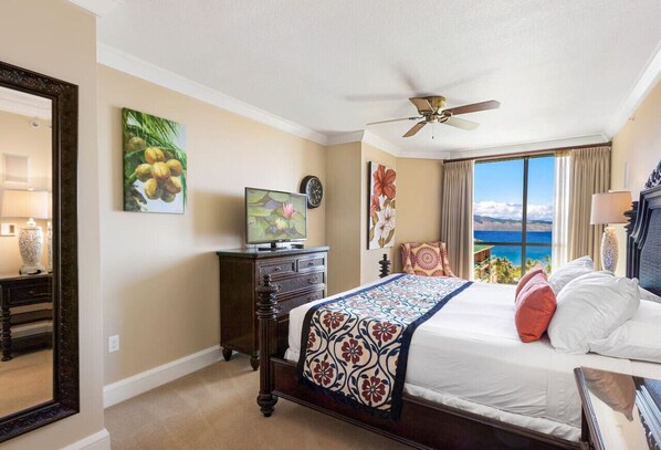 A hotel room with a large bed, a ceiling fan, a view of the sea, and artwork on the walls.