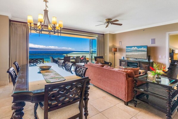 Une pièce spacieuse comprenant une table à manger, des chaises, un canapé, un téléviseur et un balcon avec vue sur l’océan.