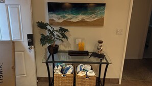 A room with a glass table, wicker baskets, a potted plant, a painting of waves, and a beach-themed sign. - Premier Condo at Cocoa Beach Towers: top floor, corner closest to Ocean + Pier (Cocoa Beach)