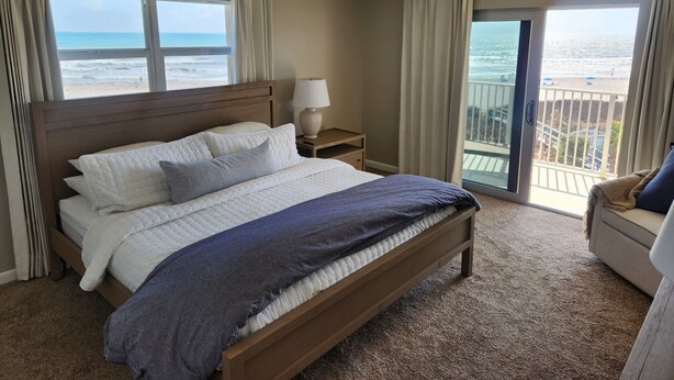 A hotel room with a large bed, a nightstand, a lamp, and a view of the ocean. - Premier Condo at Cocoa Beach Towers: top floor, corner closest to Ocean + Pier (Cocoa Beach)