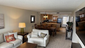A living room with a sofa, coffee table, and a dining area with chairs and a table. There is a kitchen in the background with a refrigerator and a microwave. - Premier Condo at Cocoa Beach Towers: top floor, corner closest to Ocean + Pier (Cocoa Beach)