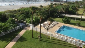 A pool area with a view of the beach and ocean, surrounded by palm trees and a white fence. - Premier Condo at Cocoa Beach Towers: top floor, corner closest to Ocean + Pier (Cocoa Beach)