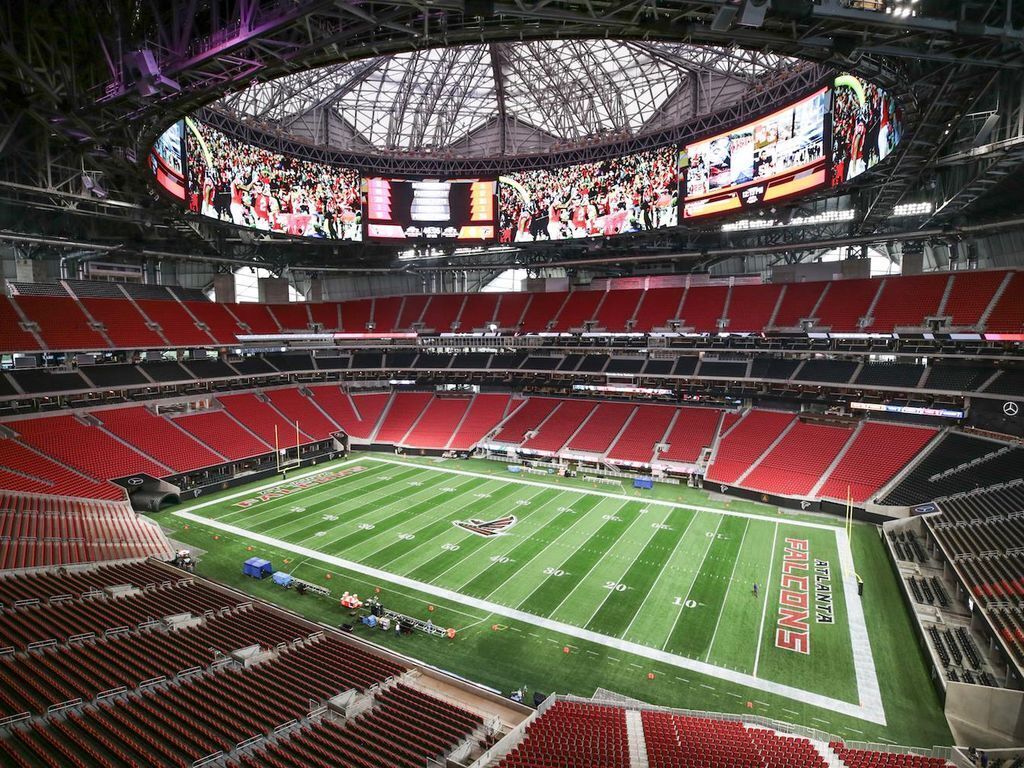 A large stadium with multiple tiers of seating, a central field marked with yard lines, and a large overhead screen displaying images.