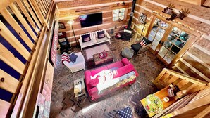 Interior de una cabaña de troncos con techo alto, chimenea, televisor y una zona de estar acogedora con un sofá rojo y un reposapiés estampado.