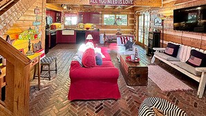 Interior espacioso de una cabaña de troncos con un sofá rojo central, piso con patrón de espiga, escalera de madera y un balcón con un letrero que dice “ALL YOU NEED IS LOVE”.