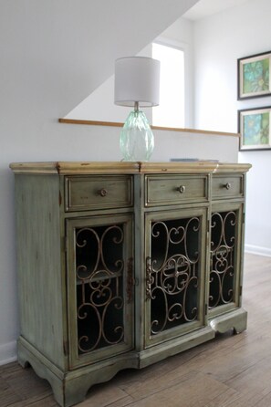 Un mueble verde de madera con detalles metálicos decorativos, un florero de vidrio con una pantalla blanca y cuadros en la pared.