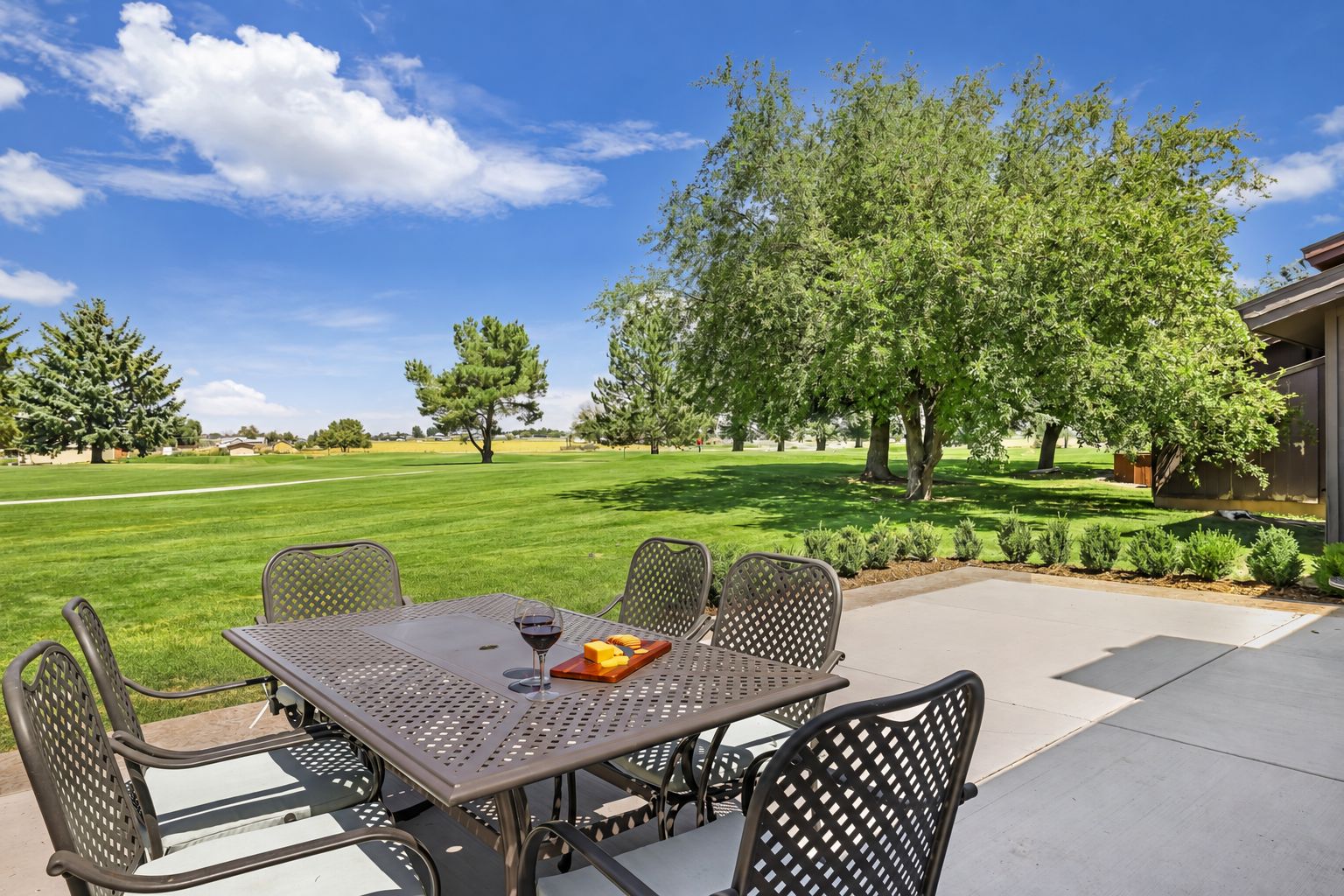 An outdoor patio with a table and chairs, a glass of red wine, and a view of a grassy field and trees.