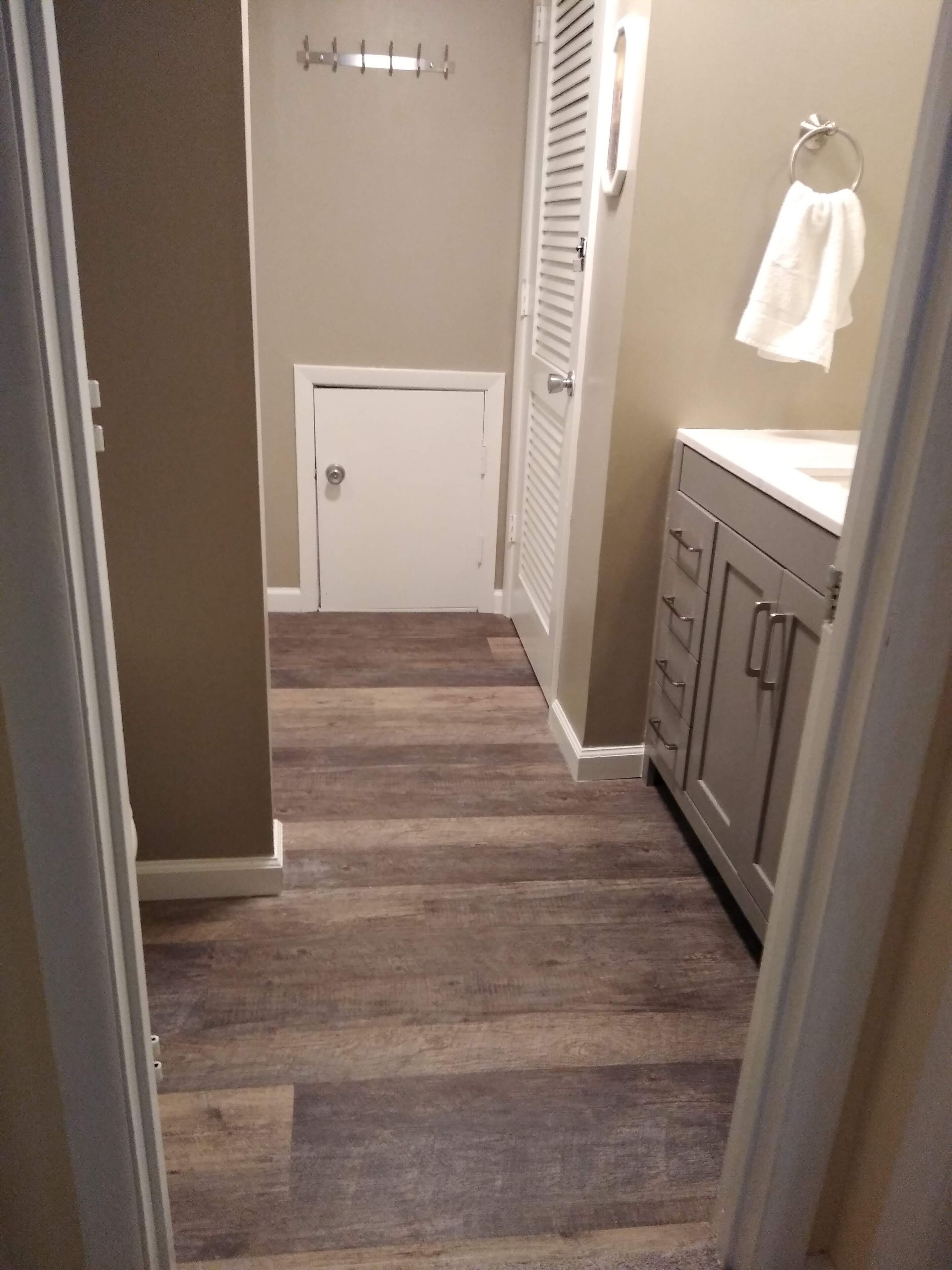 A narrow hallway with a wooden floor, a white door at the end, and a bathroom with a sink and towel rack.