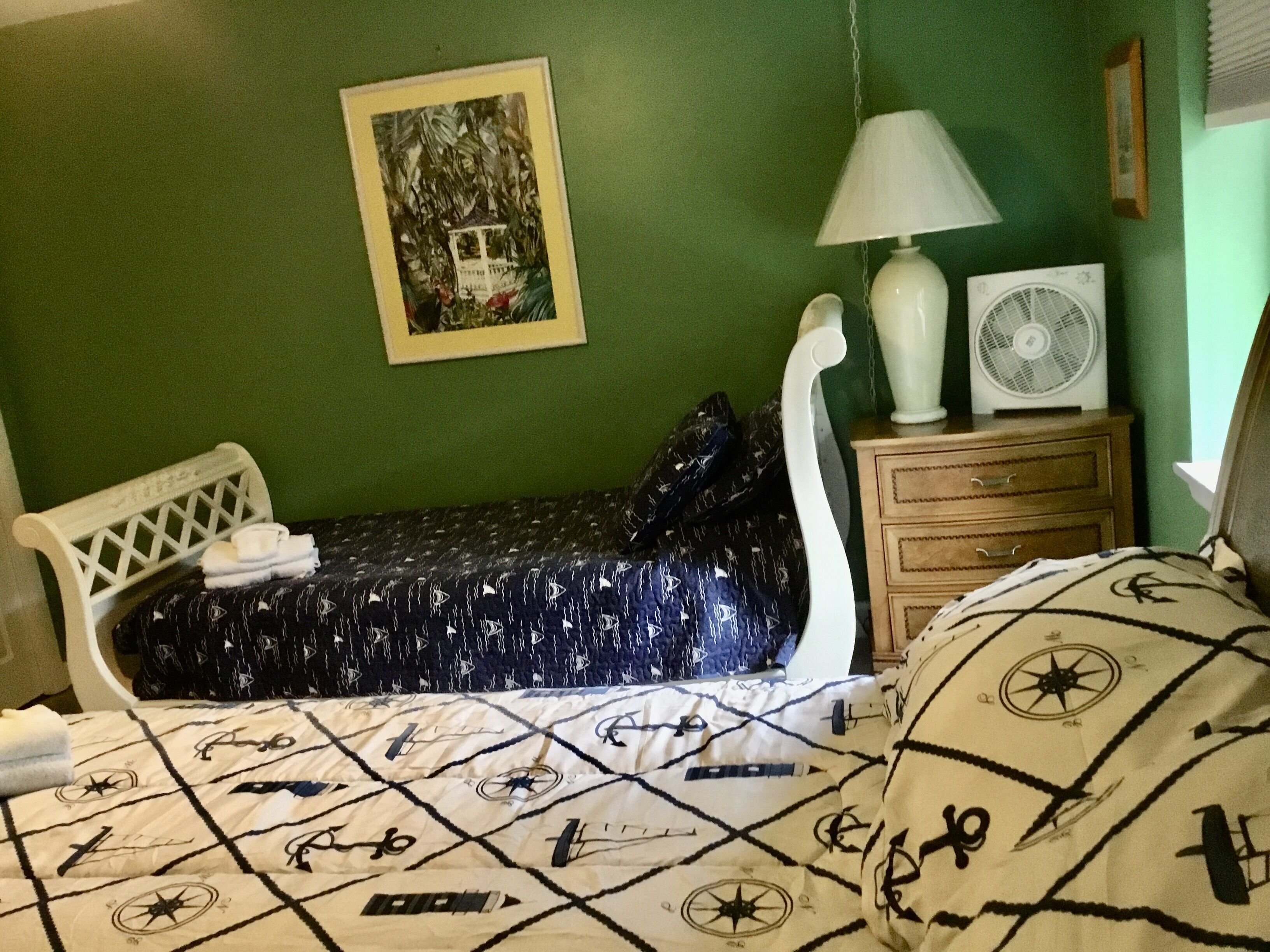 A bedroom with a bed, a bedside table with a lamp, a fan, and a framed picture on the wall.
