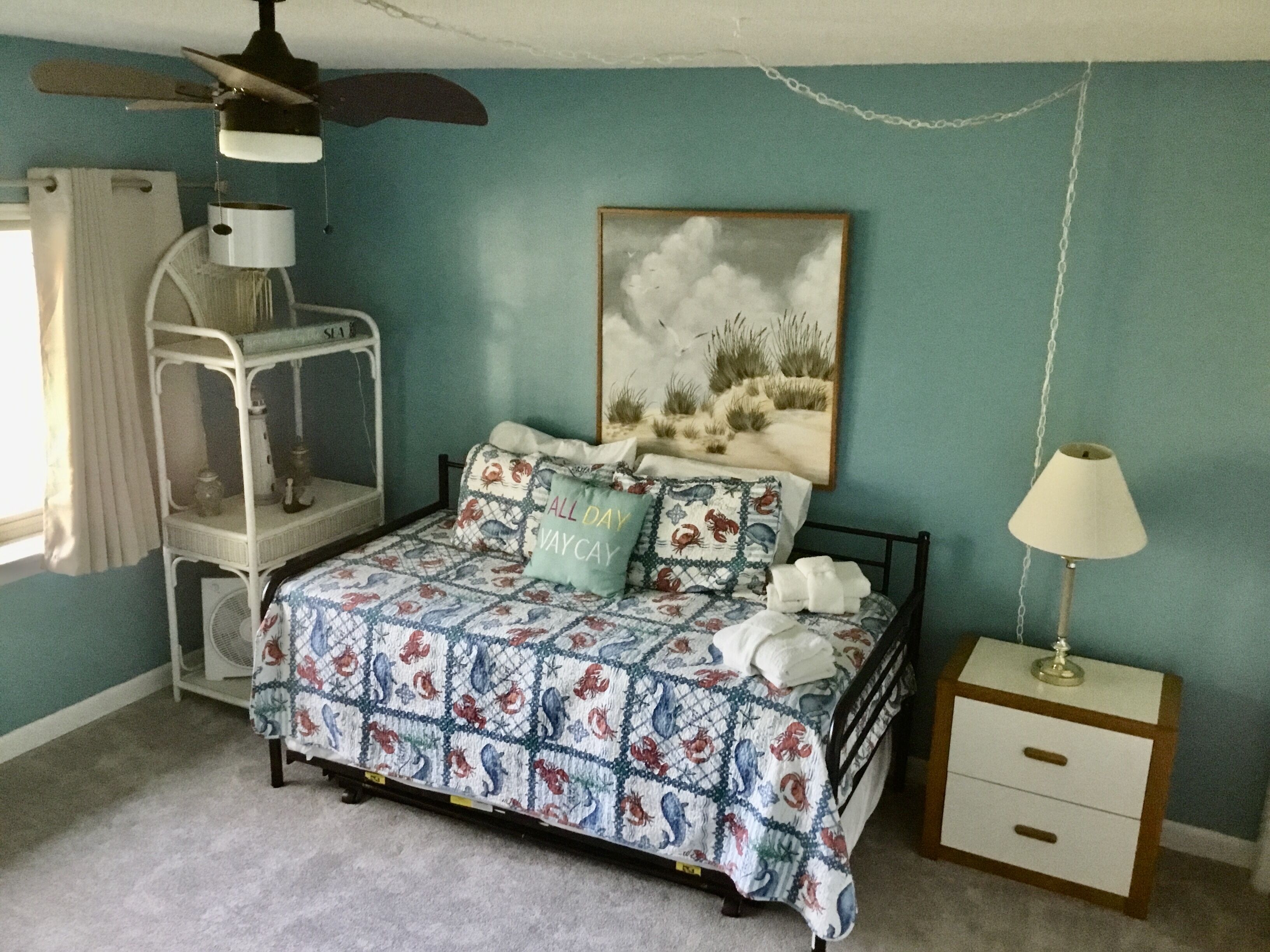 Un dormitorio con cama, mesita de noche, ventilador de techo, lámpara, estante y un cuadro en la pared.