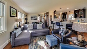 En moderne stue med en grå hjørnesofa, en sort læderstol og en lille skammel. Der er et køkkenområde med en bar og et spisebord.