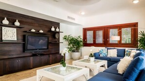 A living room with a dark wood entertainment unit, a flat-screen TV, a blue sofa, and a coffee table with a glass top. - WALKING DISTANCE TO THE BEACH (Casa de Campo)