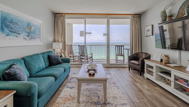 A living room with a teal sofa, a coffee table, a TV, and a view of the ocean through large sliding glass doors.