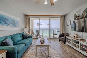 A living room with a teal sofa, a coffee table, a TV, and a view of the ocean through large sliding glass doors. - MAJESTIC BEACH RESORT-Right on the BEACH! 10% Early Booking Discounts for 2026!! (Panama City Beach)