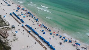A beach with rows of blue umbrellas and sun loungers, a sandy shore, and the ocean in the background.