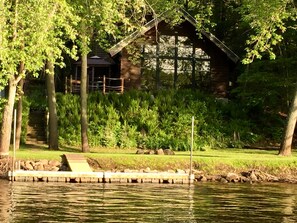 Una cabaña de madera con un muelle que se adentra en un lago tranquilo, rodeada de vegetación exuberante y árboles.