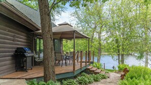 Une terrasse en bois avec des meubles d’extérieur, un barbecue et un espace salon couvert donnant sur un lac.