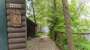 Une cabane en bois avec une pancarte accueillante, un chemin de gravier et une végétation luxuriante tout autour.
