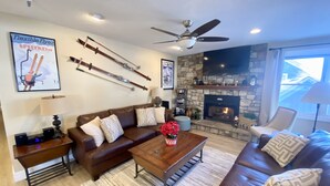 A living room with a brown leather sofa, a wooden coffee table with a vase of red flowers, a stone fireplace, and a ceiling fan. - Luxury Heart Of Old Town 3-Bdrm+ HOT TUB Walk To Skiing Chairlift Dining Main St (Park City)