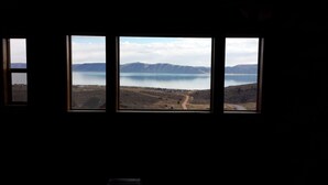 Ein Zimmer mit drei Fenstern und Blick auf einen See, Berge und eine unbefestigte Straße.