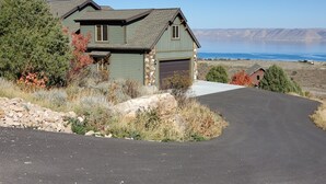 Ein zweistöckiges Haus mit Natursteinfundament und grüner Außenverkleidung, eine Auffahrt, die zu einer Garage führt, und Blick auf einen See und sanfte Hügel in der Ferne.
