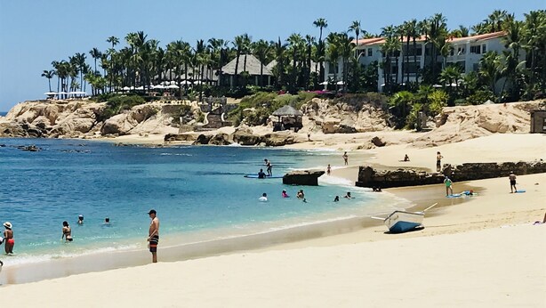 A beach with clear blue water, people swimming and sunbathing, and buildings with palm trees in the background. - Amazing Cabo Villa-Beautiful Palmilla-Near Beach (San Jose del Cabo)
