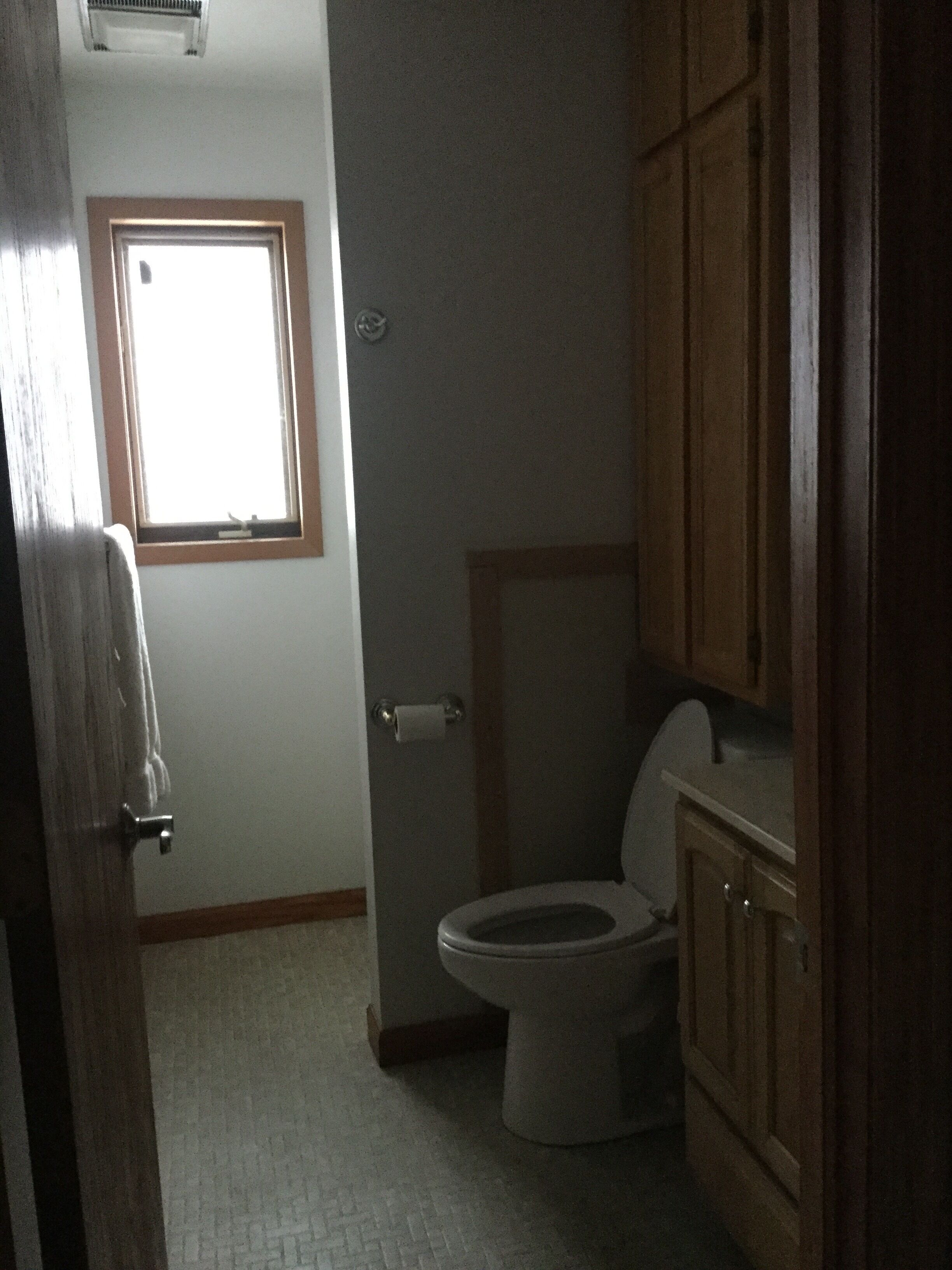 A bathroom with a toilet, wooden cabinets, a window, and a door.