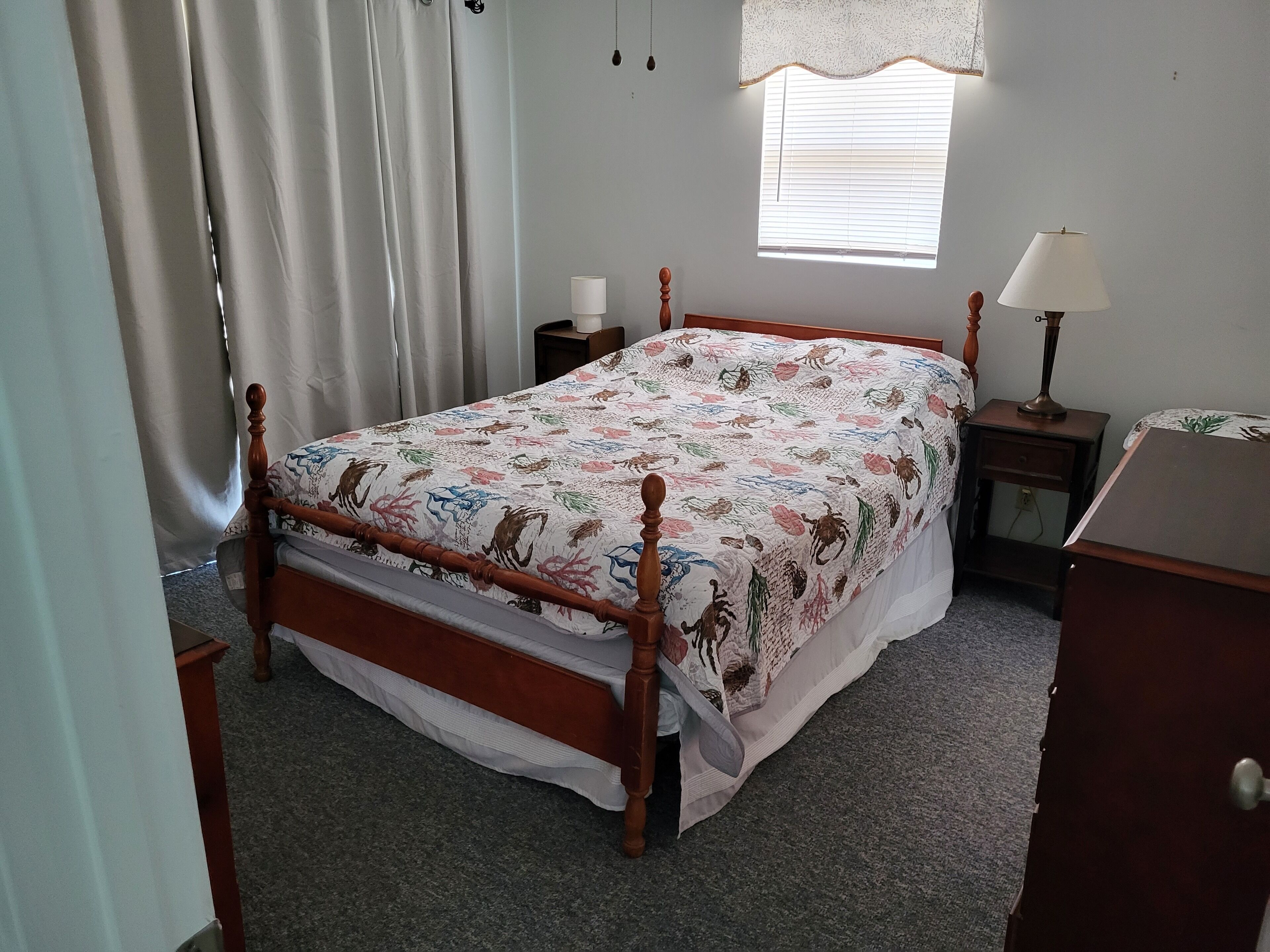 Un dormitorio con cama de madera, una ventana con persianas, una mesita de noche con lámpara y un tocador.