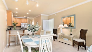 A dining area with a white table, chairs, and a vase of flowers. There is a kitchen in the background with wooden cabinets and a refrigerator. - Quiet 3BR Oasis in the Middle of Downtown Ocean City, Close to the Boardwalk. (Ocean City)