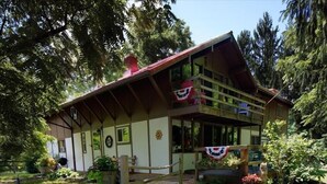A two-story wooden cabin with a balcony, surrounded by lush greenery.