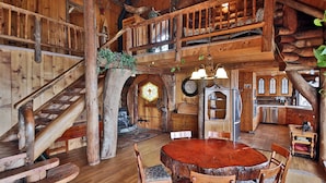 Un intérieur rustique avec des éléments en bois, notamment un escalier en bois, un coin repas avec une table ronde et des chaises, et une cuisine dotée de meubles en bois et d’une grande fenêtre.