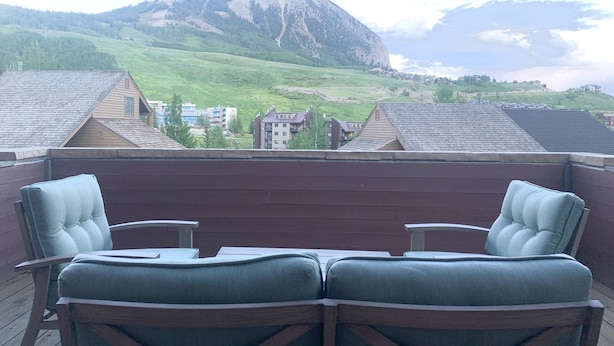 A balcony with two cushioned chairs and a table, overlooking a mountainous landscape with buildings in the valley.