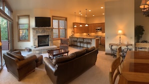 A spacious living room with a stone fireplace, leather sofas, and a television. Adjacent is a kitchen with wooden cabinets and pendant lighting.