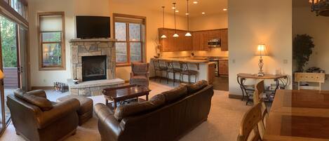 A spacious living room with a stone fireplace, leather sofas, and a television. Adjacent is a kitchen with wooden cabinets and pendant lighting.