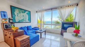 A living room with a blue sofa, a wooden cabinet, a coffee table, a TV, and a view of the ocean. - One Bedroom Beachfront Efficiency Apartment (Akumal)