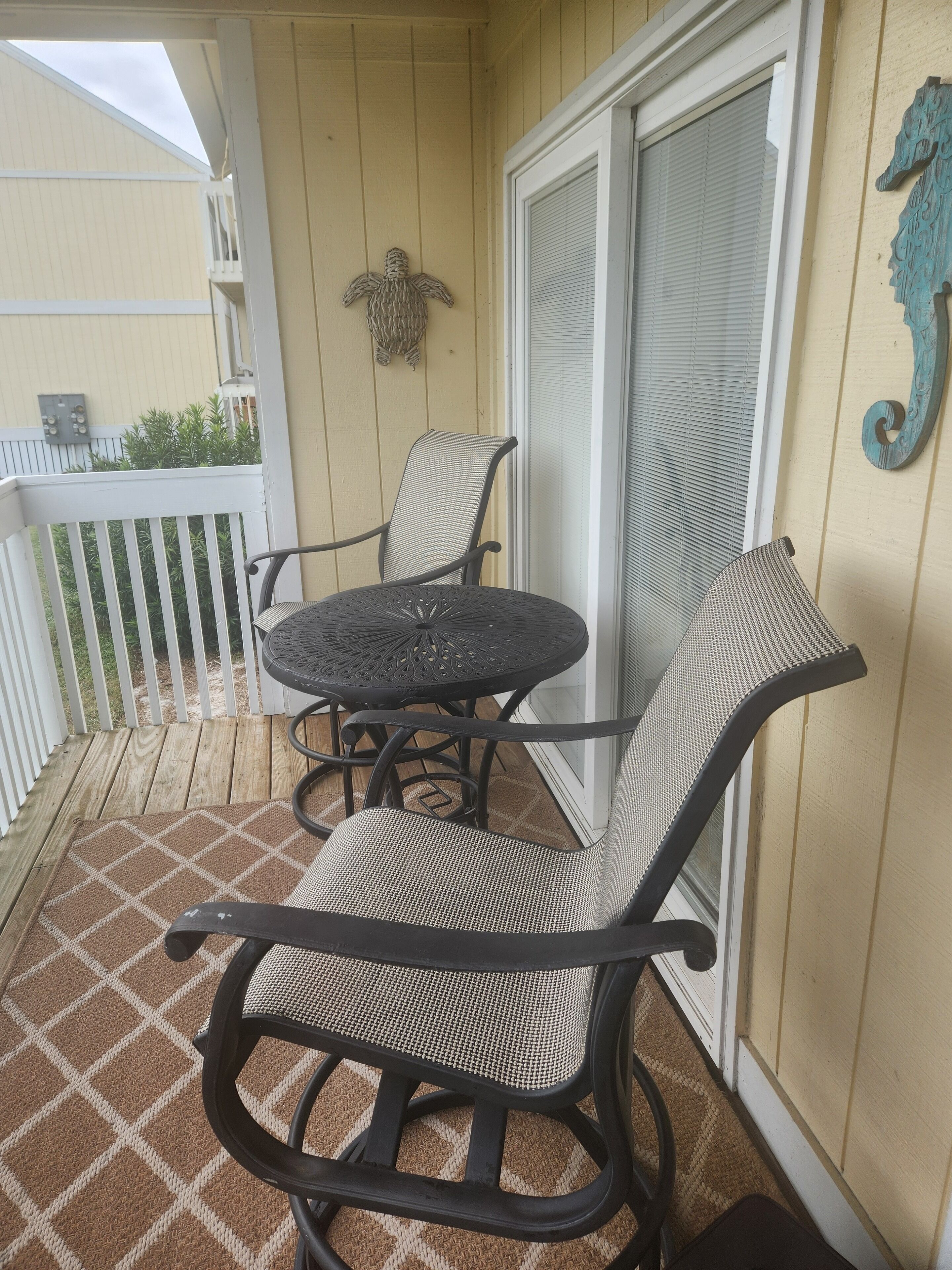 A covered outdoor seating area with a table and two chairs, a white railing, and a decorative wall piece.