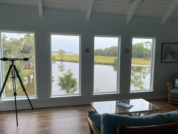 Habitación con amplias ventanas con vistas a una masa de agua, un trípode y una zona de estar con una mesa.