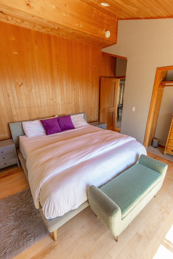 A bedroom with a wooden ceiling, a bed with white and purple bedding, a green bench, and a wooden floor.