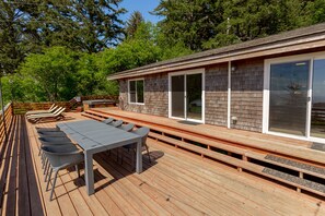 Una terraza de madera con una mesa y sillas, un porche cubierto y un edificio con ventanas amplias y una puerta corrediza.
