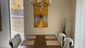 Un comedor con una mesa de madera, cuatro sillas, una araña, un cuadro y un jarrón con ramas.