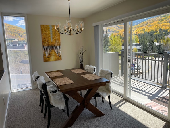 A dining area with a large table, chairs, and a chandelier. There is a painting on the wall and a view of a mountain landscape through large windows.