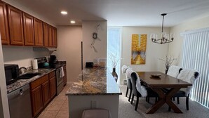 Una cocina moderna con armarios de madera, encimeras de granito y horno integrado. Hay un comedor con mesa y sillas de madera, y una araña colgante.