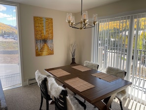 A dining area with a wooden table, black chairs, and a chandelier. There is a painting on the wall and a view of trees through the window.
