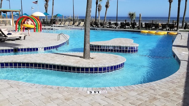 A pool area with multiple water features, including a central pool and a smaller circular pool, surrounded by palm trees and lounge chairs. - A SLICE OF PARADISE! - Direct Oceanfront condo (Myrtle Beach)