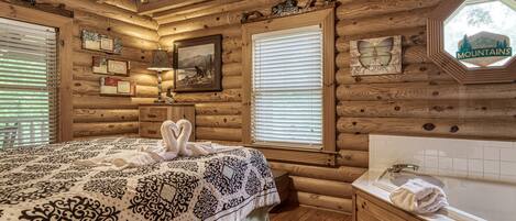 A log cabin bedroom with a bed, a vanity with a bathtub, and a window with blinds.