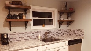 Una cocina con armarios blancos, una encimera de granito, un lavamanos de acero inoxidable y un lavavajillas. Hay dos estantes de madera sobre el lavamanos, uno con una cafetera y el otro con una botella y un tarro.
