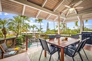 Un espacio comedor al aire libre con techo, que incluye una mesa y sillas de madera, con vista a una piscina y abundante vegetación.