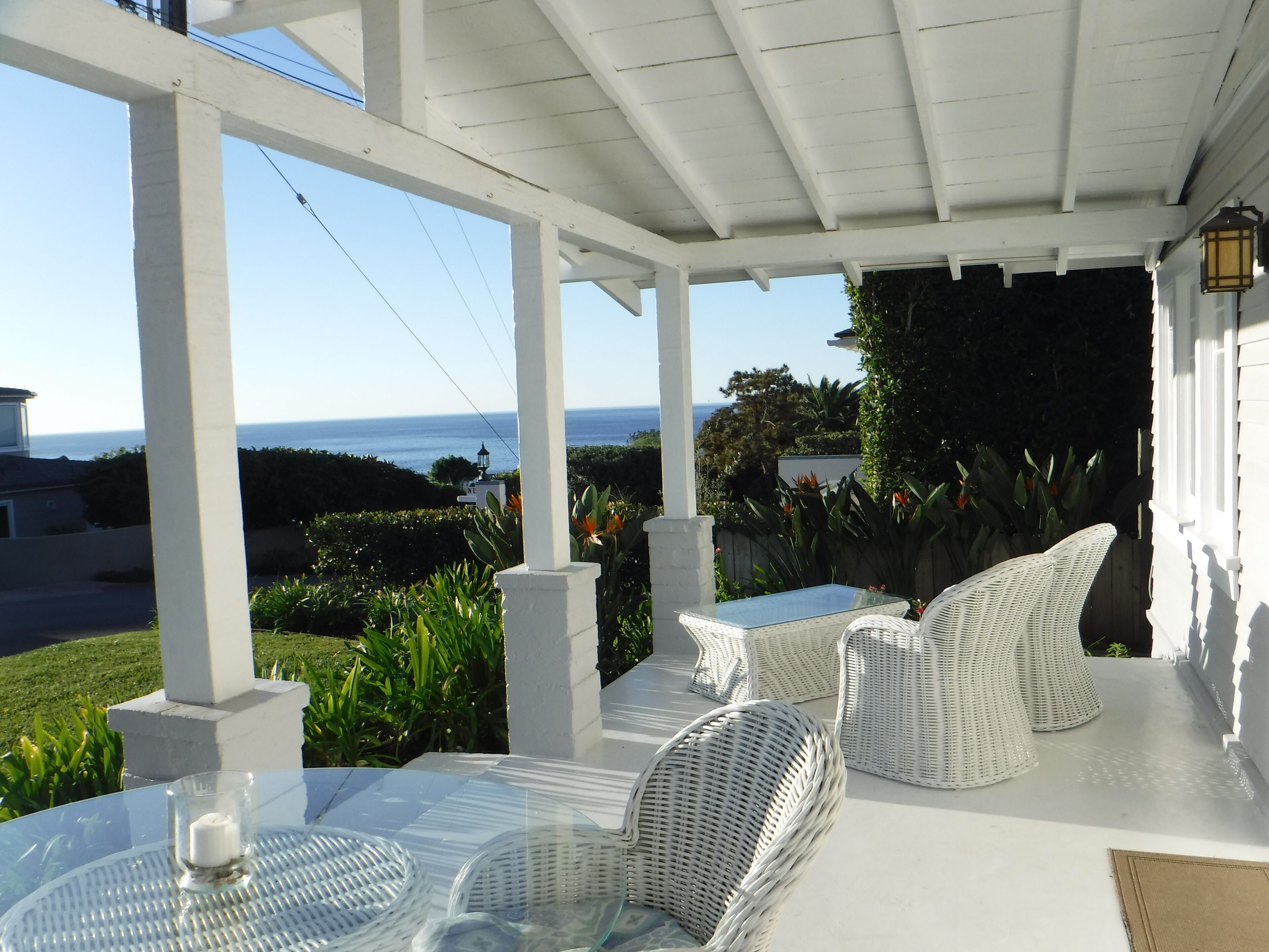 A covered patio with white wicker furniture, a glass table, and a view of the ocean.